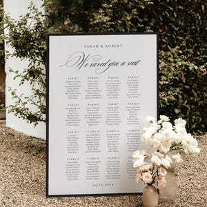 May include: A wedding seating chart with a black frame, featuring the names of guests organized by table. The text "Sarah & Robert" and "We saved you a seat" are displayed in elegant script. A vase of white flowers sits at the bottom.