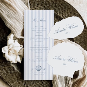 May include: A wedding menu and seashell-shaped place cards with the name "Amelia Wilson" and "Table 2" printed on them. The menu features a blue and white striped design with the text "The Menu". A white flower and fabric are also present.