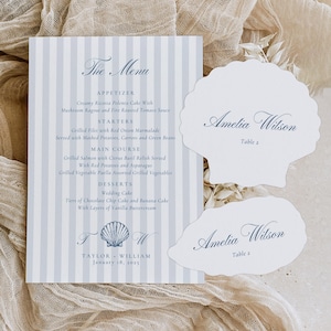 Pode incluir: Um menu de casamento e dois cartões de lugar em forma de concha. O menu apresenta um fundo listrado azul e branco com uma escrita elegante. Os cartões de lugar exibem o nome "Amelia Wilson" em uma fonte correspondente.