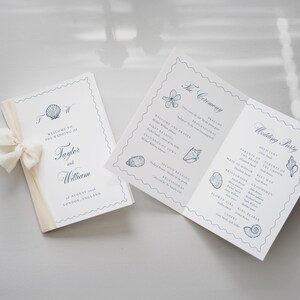 May include: A folded wedding program and a matching card featuring blue seashell illustrations, navy blue text, and a cream-colored ribbon. The text includes "Welcome to the Wedding of Taylor and William," "The Ceremony," and "Wedding Party."