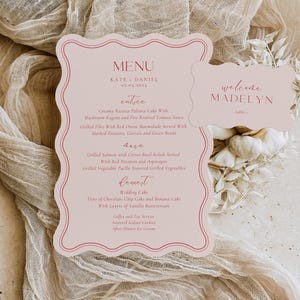 Pode incluir: Um cartão de menu de casamento rosa claro com uma borda ondulada, com os nomes "Kate & Daniel" e um menu detalhado. Um cartão menor correspondente diz "welcome MADELYN". Os cartões estão em um fundo de tecido bege.
