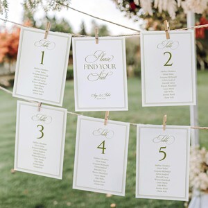 May include: Five white rectangular wedding seating chart cards with olive green text and borders, hanging from a twine string with wooden clips. The cards display the text "Table 1" through "Table 5" and "Please FIND YOUR SEAT" with names.