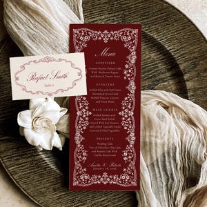 May include: A burgundy menu card and place card with white floral border designs. The menu lists appetizers, starters, main courses, and desserts. The place card reads "Rafael Smith" and "Table 6". The menu is on a textured, dark plate.
