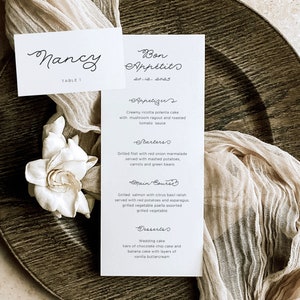May include: A white card with the word "Nancy" written in cursive and the words "Table 1" printed below. A white card with the words "Bon Appétit" written in cursive and the date "20.12.2025" printed below. The cards are on a brown wooden plate with a white cloth napkin.
