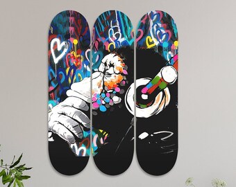 Op de afbeelding: Drie zwarte skateboard decks met een kleurrijk graffiti ontwerp met een persoon die een microfoon vasthoudt. De graffiti bevat harten, sterren en andere abstracte vormen.
