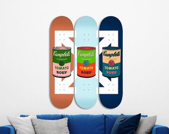 Op de afbeelding: Drie skateboards met een wit pijlontwerp en een Campbell's Tomato Soup-blikje-afbeelding. De skateboards zijn in een rij op een witte muur geplaatst, met het meest linkse skateboard oranje, het middelste skateboard blauw en het meest rechtse skateboard donkerblauw.