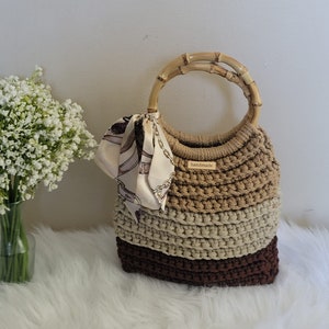 May include: A crocheted tote bag with a brown, beige, and cream striped design. The bag has a bamboo handle and a small "Handmade" tag attached to the front.