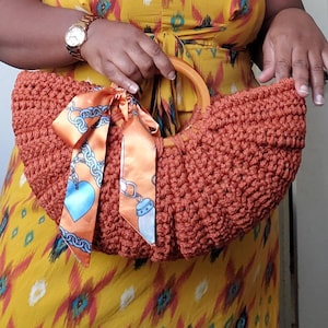 May include: A crocheted handbag with a wooden handle and a colorful scarf tied around the handle. The bag is a burnt orange color and has a textured, crocheted design.