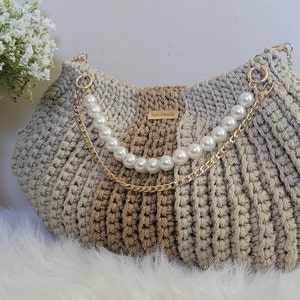 May include: A crocheted handbag with a beige and gray color gradient. The bag has a gold chain strap with pearl accents and a small tag that reads "handmade".