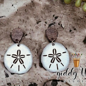 May include: A pair of wooden earrings with a white sand dollar design. The earrings are brown and have a silver hook.