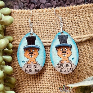 May include: A pair of wooden earrings featuring a cartoon groundhog wearing a top hat. The earrings are oval shaped and painted in shades of blue, brown, and white. The text "Ground Hog Day" is written on the earrings.