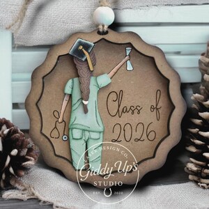 K&ouml;nnte beinhalten: Holzornament mit einer graduierenden Krankenschwester in OP-Kleidung, die einen Becher h&auml;lt und eine Abschlusskappe tr&auml;gt. Das Ornament tr&auml;gt die Aufschrift "Class of 2026" und stammt von Giddy Up's Studio. Das Design wurde von Stacey Hutton gemalt.