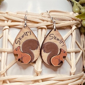 May include: A pair of wooden dangle earrings with a squirrel design. The earrings are brown and feature a squirrel with a bushy tail. The earrings are labeled "Squirly" on the back.