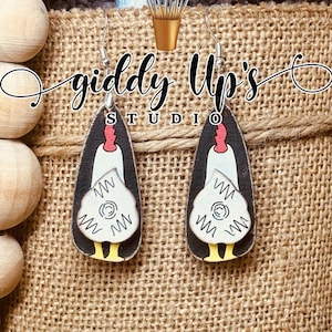 May include: A pair of wooden earrings shaped like chickens with white and black details. The earrings are hanging from a burlap background with the text "giddy Up's Studio".