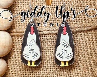 May include: A pair of wooden earrings shaped like chickens with white and black details. The earrings are hanging from a burlap background with the text "giddy Up's Studio".
