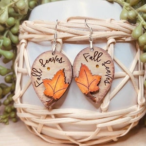May include: A pair of wooden earrings with a fall leaves design. The earrings are shaped like leaves and have the words "Fall Leaves" written on them. The leaves are painted in shades of orange, yellow, and brown.