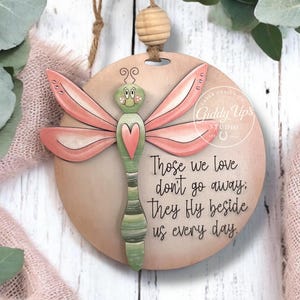 May include: Wooden dragonfly ornament with pink wings and a green body, featuring the text "Those we love don't go away: They fly beside us every day." Includes a jute string for hanging. SVG digital download.