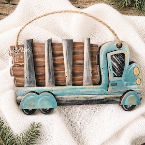 May include: A wooden truck ornament featuring a teal cab with yellow headlights and a flatbed carrying brown logs. The truck has a distressed finish and a jute twine hanger attached to the top. The text Giddy Up's Studio is visible on the cab.