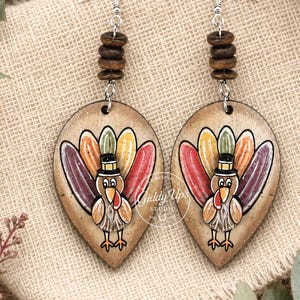 May include: Teardrop-shaped wooden earrings featuring a cartoon turkey illustration with a black top hat and colorful feathers. The earrings include silver-toned metal hooks and a stack of three small brown wooden beads above each turkey pendant.