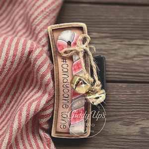 May include: A wooden candy cane ornament with a red and white striped design, tied with twine and gold bells. The ornament has the text "#candycane lane" and "Giddy Up's Studio" on it. The background includes a red and white striped fabric.
