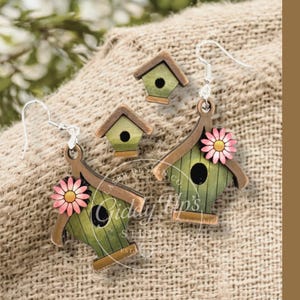 May include: Wooden birdhouse earrings with a matching birdhouse charm. The earrings and charm are painted green with brown trim and a pink flower accent. The earrings have silver hooks. The text on the image says "SVG DIGITAL DOWNLOAD. NO PHYSICAL ITEM WILL BE SENT."