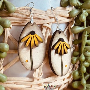 May include: A pair of wooden earrings with a yellow flower design. The earrings are oval shaped and have a silver hook.