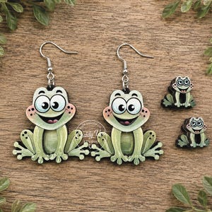May include: A pair of dangling earrings and two smaller matching charms, each featuring a cartoon frog with large eyes, a wide smile, and light green skin. The text on the right side reads: SVG DIGITAL DOWNLOAD. NO PHYSICAL ITEM WILL BE SENT.