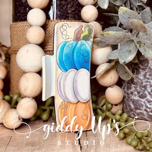 May include: A wooden sign with a stack of three pumpkins painted in blue, white, and orange. The sign is decorated with a brown stem and green leaves. The sign reads "giddy up's studio".