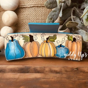 May include: A wooden hair clip featuring a hand-painted design of pumpkins in shades of blue, orange, and yellow. The clip has a teal top and is decorated with green leaves and vines. The words "giddy Up's STUDIO" are written on the clip.