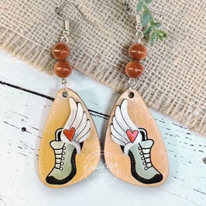 May include: Dangle earrings with a light brown wooden teardrop shape, featuring a painted design of a boot with wings and a heart. The earrings have silver hooks and two brown beads.