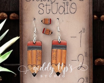 May include: A pair of pencil-shaped earrings and two small pencil-shaped studs displayed on a wooden stand labeled Giddy Up's Studio. The stand features a printed ruler scale from 1 to 3 inches. The text PENCIL EARRING SET is visible on the right.
