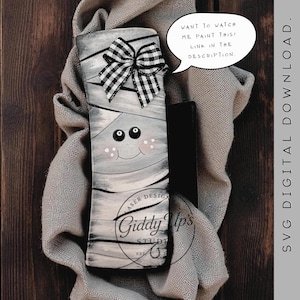 May include: A painted wooden mummy decoration with a black and white checkered bow. The mummy has a smiling face with rosy cheeks and is wrapped in gray bandages. Text on the image says, "Want to watch me paint this! Link in the description."