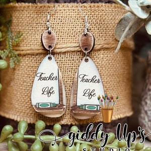 May include: Wooden Teacher Life earrings with a sailboat design. The earrings are white with brown accents and feature the words "Teacher Life" and a small illustration of a book and pencil. The earrings are hanging from silver hooks.