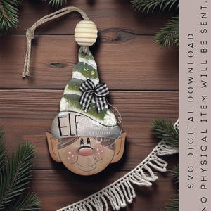 May include: Wooden elf Christmas ornament. The elf has a painted face with rosy cheeks and a red nose. The elf's hat is green and white with a black and white checkered bow. The word 'ELF' is painted on the elf's face. The ornament hangs from a string with a wooden bead.