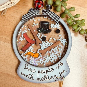 May include: A white snowman ornament with a black top hat, red scarf, and orange carrot nose. The ornament is filled with white beads and glitter. The text "Some people are worth melting for." is written on the bottom of the ornament. The ornament is made by Giddy Up's Studio.