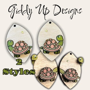 May include: Two styles of wooden earrings featuring a turtle design. The turtle is green with a pink shell and is outlined in black. The earrings are shaped like a teardrop and have a gold post.