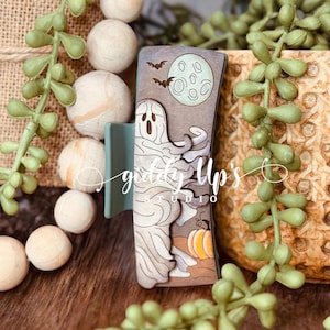 May include: A wooden hair clip with a green base and a design of a ghost flying in front of a full moon with bats. The moon is green and the ghost is white. The background is brown with a pumpkin in the bottom right corner.