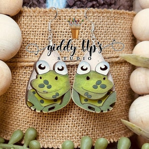 Puede incluir: Un par de pendientes de rana verde con ojos blancos y un fondo de madera marrón. Los pendientes están hechos de madera y tienen un acabado brillante. Los pendientes están etiquetados como "giddy Up's Studio".