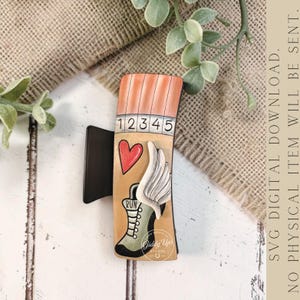 May include: A wooden hair clip featuring a running theme. The clip has a tan background with a red heart, white wings, and a green boot with the word "RUN". The top has orange stripes and the numbers 1-5. The clip is held by a black metal clasp.
