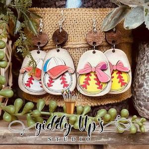 May include: A set of four wooden earrings featuring a softball design. The earrings are painted in various colors, including yellow, pink, red, blue, and white. The earrings are shaped like a softball with a baseball cap on top. The earrings are hanging from brown wooden earring hooks. The earrings are labeled "giddy Up's STUDIO".
