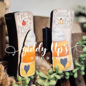May include: Two wooden candy corn decorations with painted details. One says "trick or treat" and the other says "sweet & corny". Both have a black heart and are painted in orange, yellow, and white.