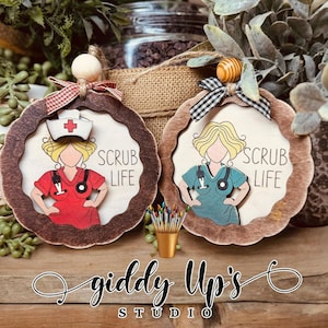 May include: Two wooden ornaments with a nurse theme. One features a red uniform and the other a teal uniform. Both have the text "SCRUB LIFE" and are decorated with a ribbon and wooden bead.