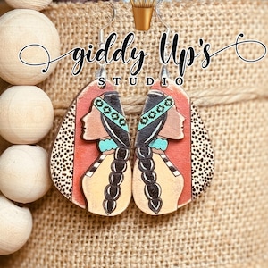 May include: A pair of Native American-inspired earrings with a red, white, and black geometric design. The earrings feature a woman's face with turquoise accents and black braids. The earrings are made of wood and have silver hooks.