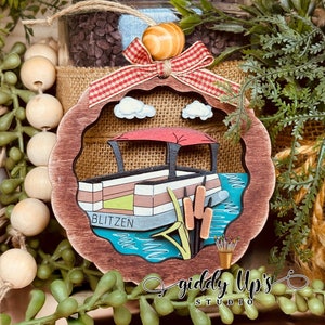 May include: A wooden Christmas bauble shaped like a boat with a red awning and the word "Blitzen" on the side. The bauble is painted in shades of brown, green, blue, and yellow. The bauble is hanging from a red and white checked ribbon.