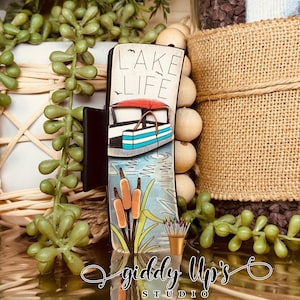 May include: A wooden sign with the text "Lake Life" painted in white. The sign features a cartoon boat with a red awning and blue and white stripes. The boat is surrounded by green reeds and blue water. The sign is decorated with a brown wooden frame and a black metal clip. The sign is made by Giddy Up's Studio.