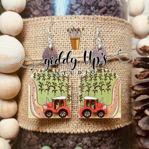 Puede incluir: Un par de pendientes de madera con un tractor rojo conduciendo por un campo de maíz. Los pendientes están pintados en verde, marrón y rojo. Los pendientes están etiquetados como "giddy Up's Studio".