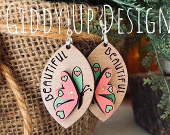 May include: A pair of wooden earrings featuring a butterfly design. The earrings are painted in shades of pink, green, and white. The word "Beautiful" is written on each earring.