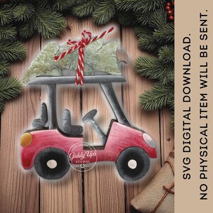 Puede incluir: Adorno navideño de madera roja con forma de carrito de golf con un árbol verde atado en la parte superior con hilo rojo y blanco. El adorno tiene ruedas negras y un faro amarillo. El texto "Giddy Up's Studio" está en el lateral. Fondo con ramas de pino.