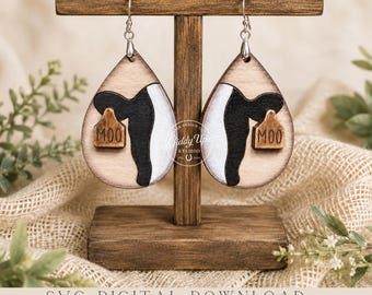 May include: Teardrop-shaped wooden earrings featuring a black and white cow print pattern. Each earring displays a small brown tag with the text MOO engraved on it. The earrings hang from silver-toned metal hooks and are displayed on a wooden stand.