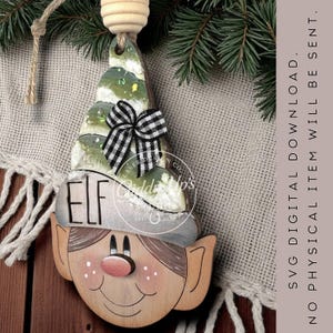 May include: Wooden elf Christmas ornament with a painted face, hat, and a green and white tree-shaped top. The ornament has a black and white checkered bow and the word "ELF" on the hat. The text "SVG DIGITAL DOWNLOAD. NO PHYSICAL ITEM WILL BE SENT." is also visible.